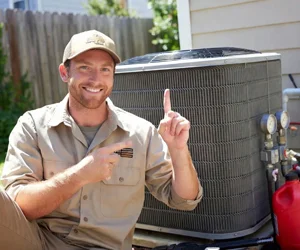 Smiling HVAC technician in uniform pointing up next to outdoor air conditioning unit in Simi Valley
